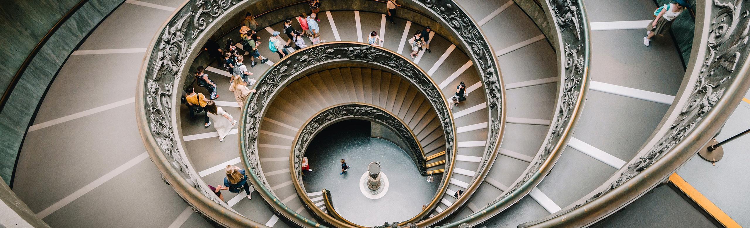 Vue en plongée d’un escalier en colimaçon monumental avec rampe décorée, rempli de visiteurs.