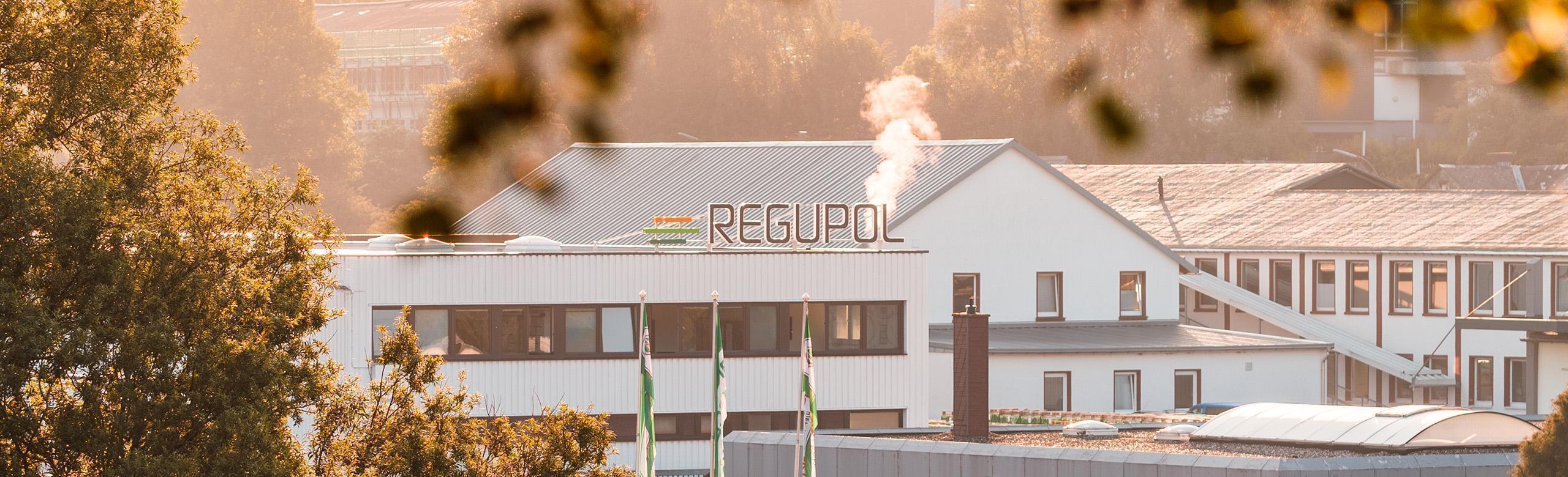 Site de production REGUPOL – usine, durabilité, environnement, industrie Vue extérieure d’un site de production REGUPOL au petit matin. Le bâtiment industriel blanc porte le logo REGUPOL sur le toit, avec de la vapeur s’échappant d’une cheminée. Des arbres et des drapeaux verts de REGUPOL sont visibles au premier plan.