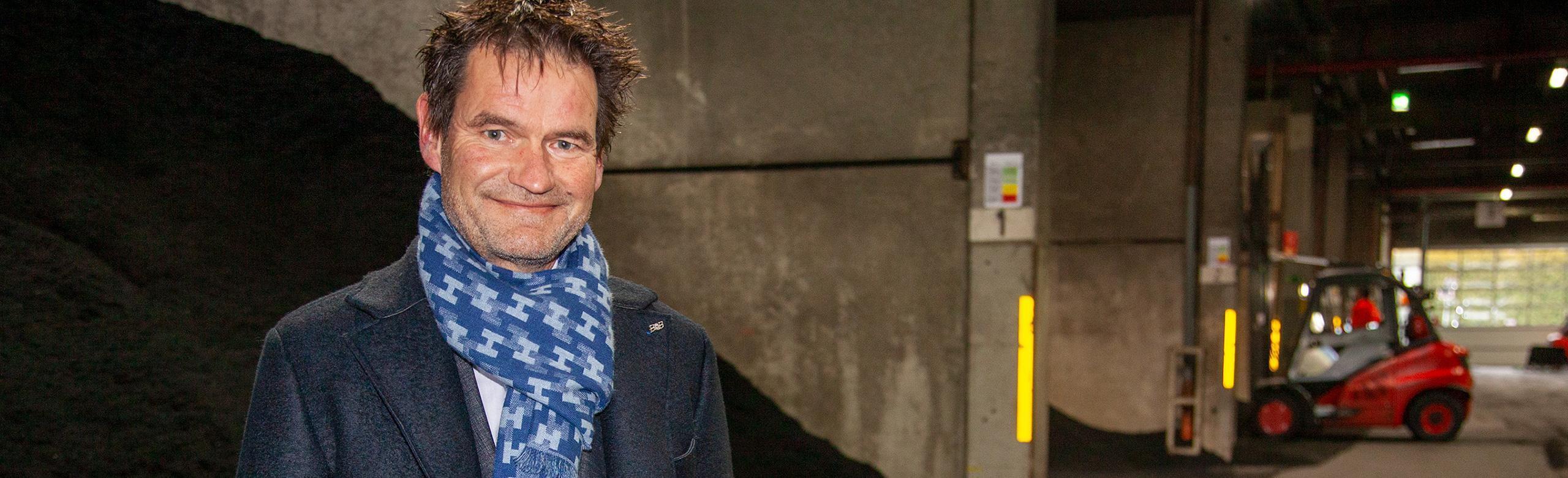 Dirigeant REGUPOL sur site – recyclage, industrie, durabilité, direction Un des dirigeants de REGUPOL se tient dans un entrepôt industriel, vêtu d’un manteau foncé et d’une écharpe bleue. À l’arrière-plan, des matériaux en vrac et un chariot élévateur rouge.