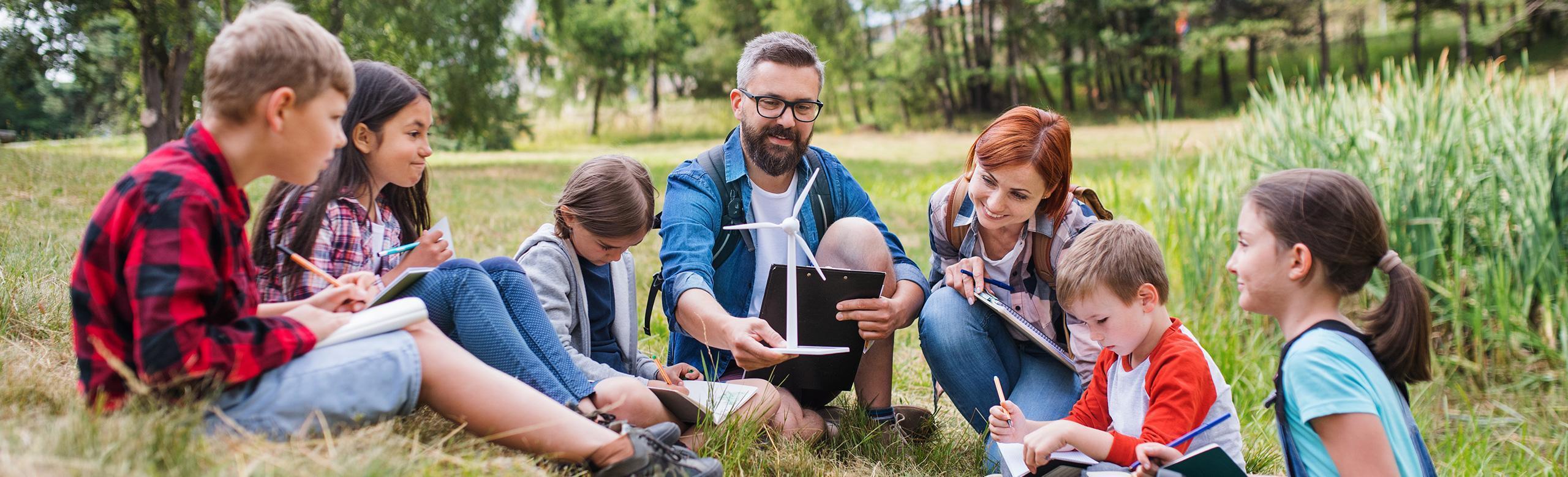 Éducation à l’environnement en plein air – enfants, nature, éolienne, durabilité Un groupe d’enfants est assis avec deux adultes en cercle sur une prairie. Ils apprennent en plein air avec des cahiers et un modèle d’éolienne. Arbres et roseaux en arrière-plan.